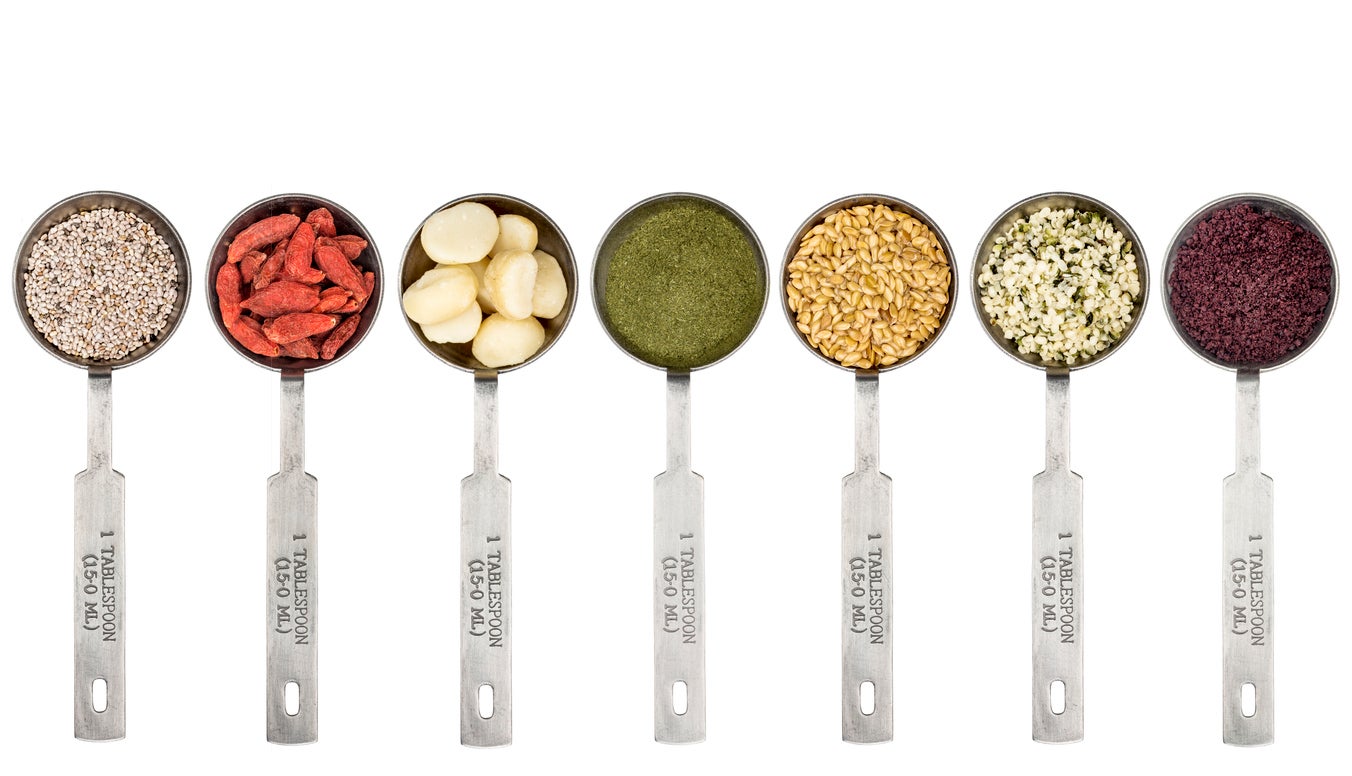 superfood abstract - white chia seeds, goji berry,macadamia nuts, barley grass powder, golden flax seeds, hemp seeds and acai berry powder - top view of  metal measuring tablespoons isolated on white