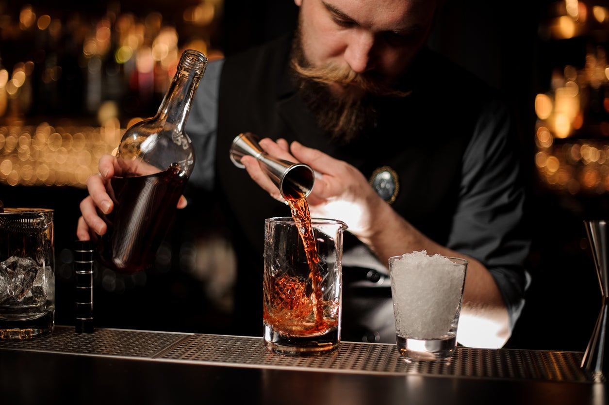 Bartender pouring a brown alcohol to the cocktail from the steel jigger to a glass on the bar counter on the blurred background