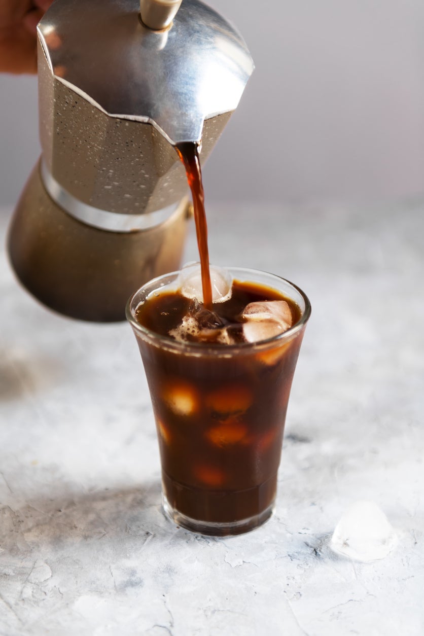 Fresh brew black coffee is pouring from mocha into a tall glass with pieces of ice on a gray dark background, close-up. Ice summer coffee concept, close up